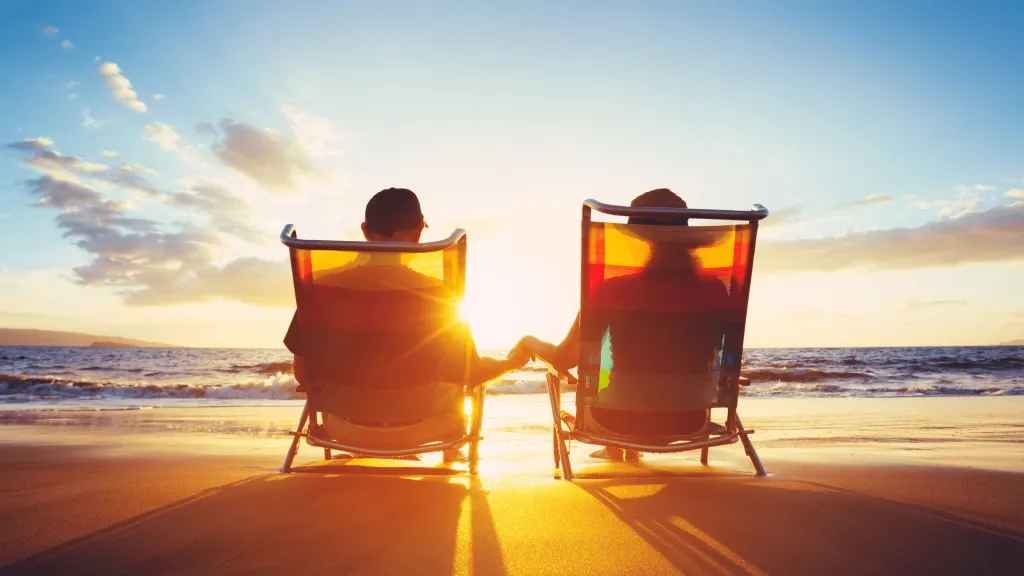 Un couple assis dans des chaises de plage se tient la main en admirant le coucher de soleil face &agrave; la mer.