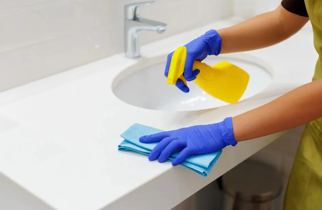 Une personne portant des gants nettoie un lavabo &agrave; l'aide d'un chiffon et d'un spray.