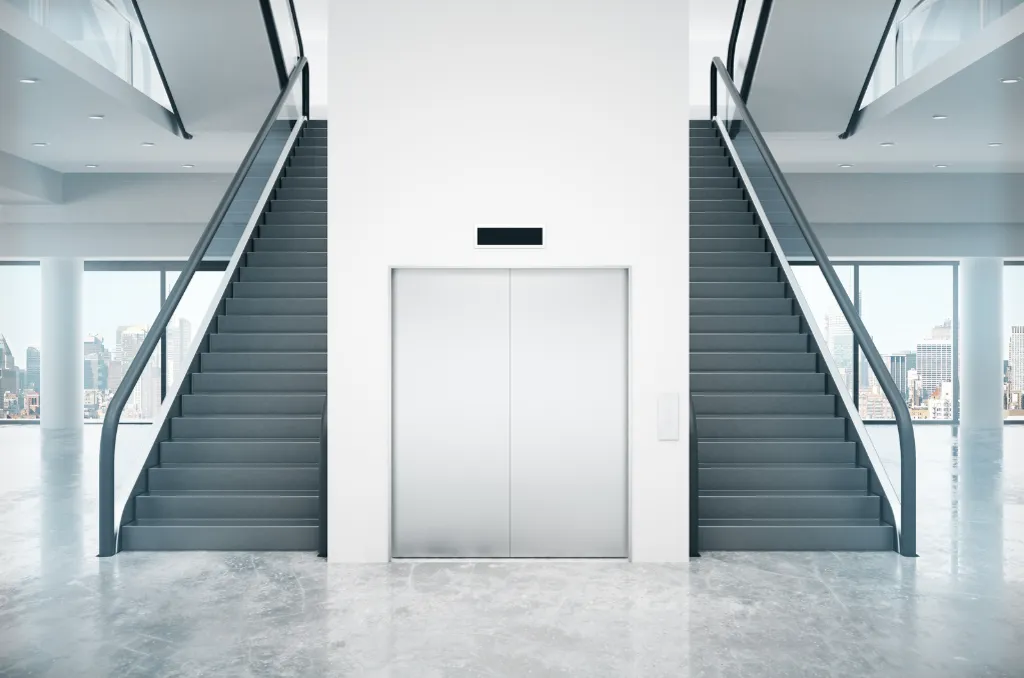 Hall d'immeuble moderne avec un ascenseur central, des escaliers m&eacute;caniques de chaque c&ocirc;t&eacute; et une grande fen&ecirc;tre offrant une vue sur la ville.